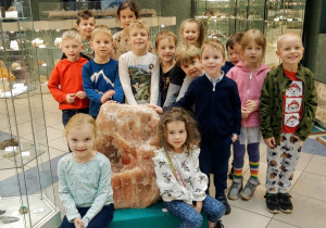 Grupa dzieci pozuje do zdjęcia w Muzeum Geologicznym w scenerii różnorodnych skał umieszczonych za szybami.